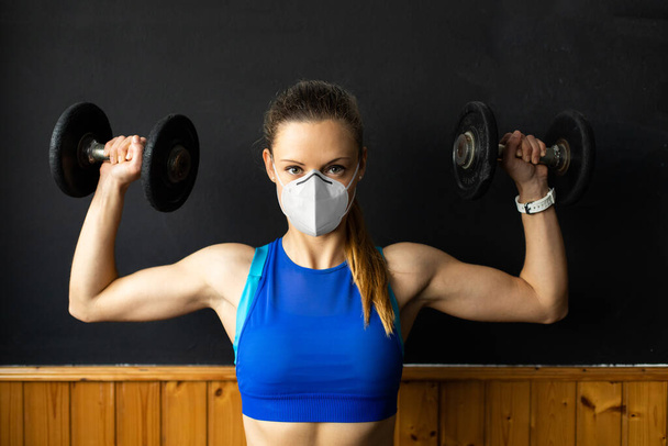 Young fit woman with n96 face mask  doing shoulder press exersice at the gym with dumbbells. Fitness strength workout under coronavirus health crisis. - Photo, image