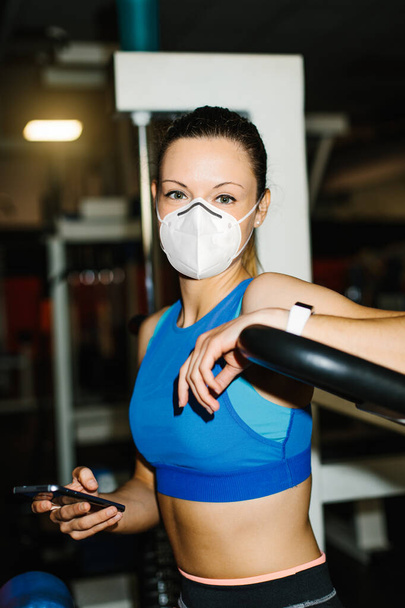 Young fitness woman working out in the gym wearing n95 face mask for protecting against Covid-19 infection. Fit girl taking a break for texting on smartphone. - 写真・画像