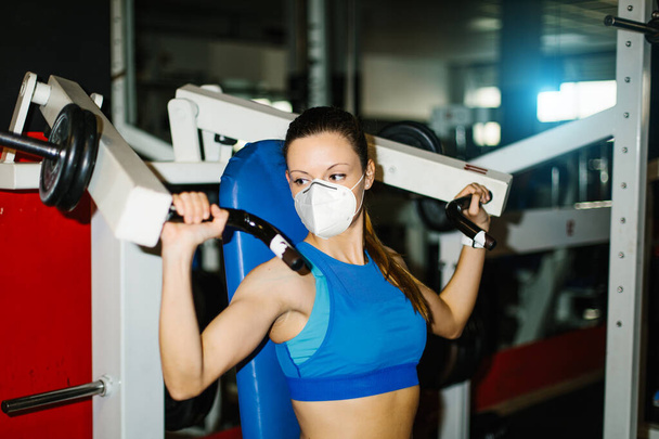 Jeune femme de fitness travaillant dans la salle de gym portant un masque facial n95 pour se protéger contre l'infection Covid-19. Exercice de machine d'épaule
. - Photo, image