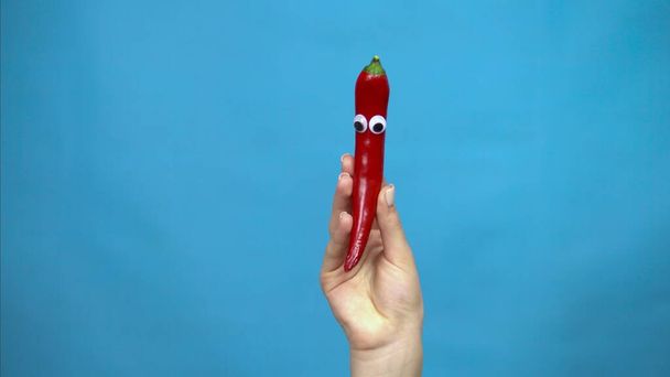 Chilli-pippuri, jolla on silmät naisen kädessä sinisellä pohjalla. Nainen käsi lähikuva. - Valokuva, kuva