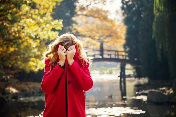 young woman hiding under red coat  in autumn city park near lake - Foto, afbeelding