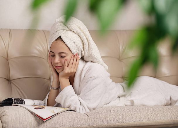 Attractive girl with towel on her head and in white bathrobe. Lies on  couch and watches a magazine, in good mood. - Φωτογραφία, εικόνα