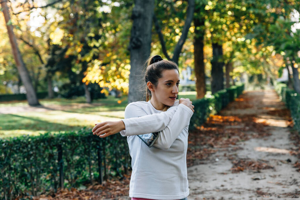 Young athlete woman stretches her left arm on a path in a leafy park with green background - Fotó, kép