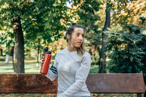 Mujer atleta sentada en un banco del parque con una botella roja con agua descansando del entrenamiento
 - Foto, Imagen