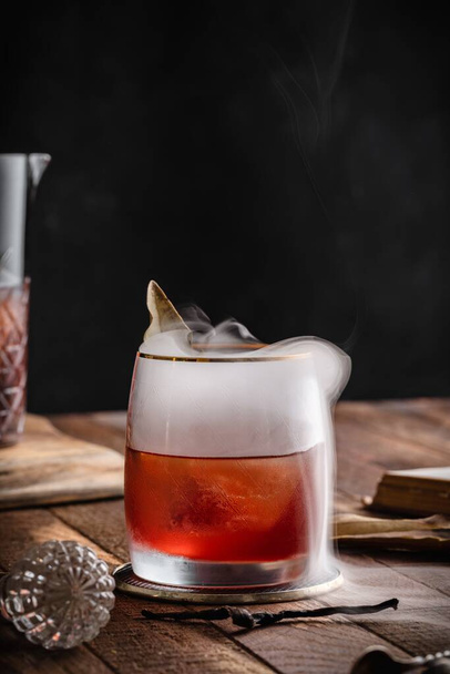 A vertical shot of a brown cocktail in a glass with smoke placed on a wooden surface - 写真・画像
