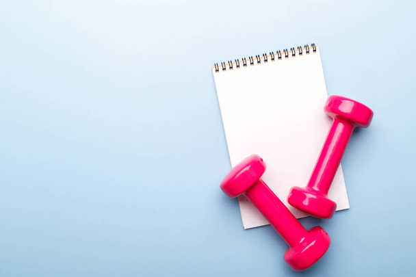 Pink dumbbells and notepad on blue background, flat lay copy space - Photo, Image