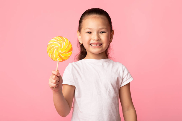 Αξιολάτρευτο κορίτσι από την Ασία χαμογελώντας και κρατώντας πολύχρωμο γλειφιτζούρι - Φωτογραφία, εικόνα