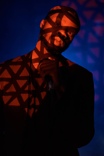 dark-skinned handsome guy in a bandana, black classic jacket and t-shirt holding a microphone in his hand,standing in the shadow  on a blue light background and looking at the camera - Foto, Bild