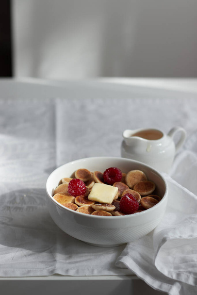 Bowl with tiny pancake cereal with raspberries, butter and honey on a white material. Trendy food. Mini cereal pancakes. Trendy breakfast during quarantine. - Foto, afbeelding