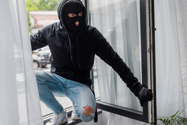 Selective focus of robber in balaclava standing on open window  - Photo, Image