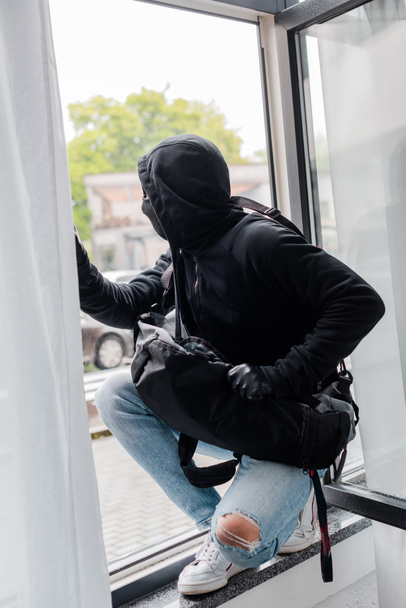 Selective focus of robber in balaclava and leather gloves holding bag while standing on window  - Фото, изображение