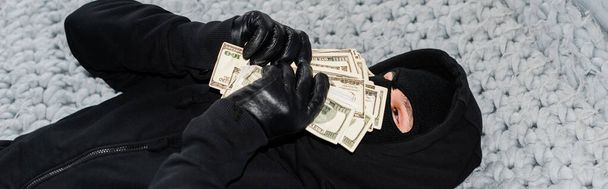Panoramic shot of thief in balaclava holding money near face on bed  - Foto, Imagem