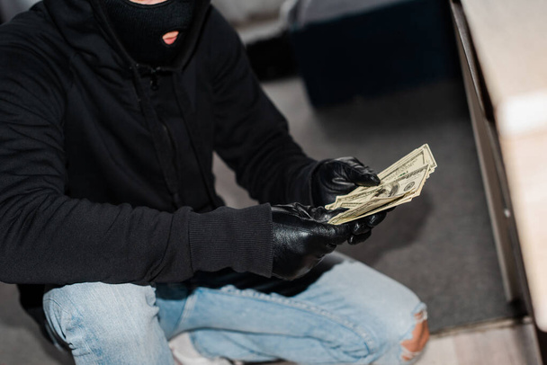 Selective focus of robber in mask and leather gloves holding dollars during theft - Photo, Image