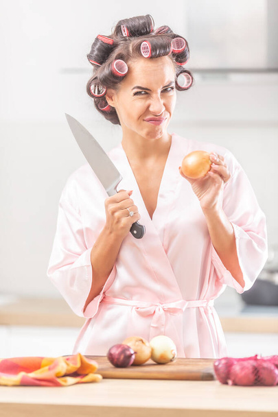 Female with hair rollers and silk pyjamas frowns upon the need to cut the onion by a knife. - Foto, immagini