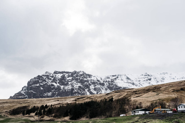 白い家と丘の上の森の近くの建設掘削機、アイスランドの岩場の雪に覆われた山の背景. - 写真・画像