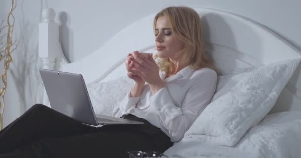 Businesswoman working on laptop enjoys a cup of hot coffee before bedtime after a long hard day. Business woman relaxes in bed after a long flight. Business concept. Side angle close up. Prores 422 - Séquence, vidéo