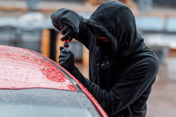 Selective focus of robber in mask and leather gloves opening door of auto with screwdriver outdoors - Foto, Imagem