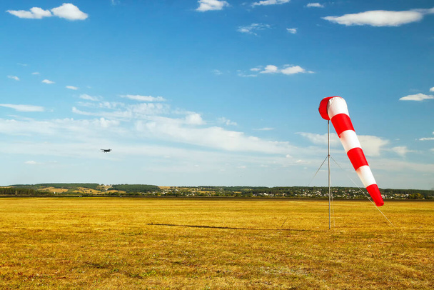 rot-weiße Windsack-Windsocke am blauen Himmel, gelbes Feld und Wolken im Hintergrund, Silhouette eines Oldtimer-Flugzeugs am Himmel - Foto, Bild