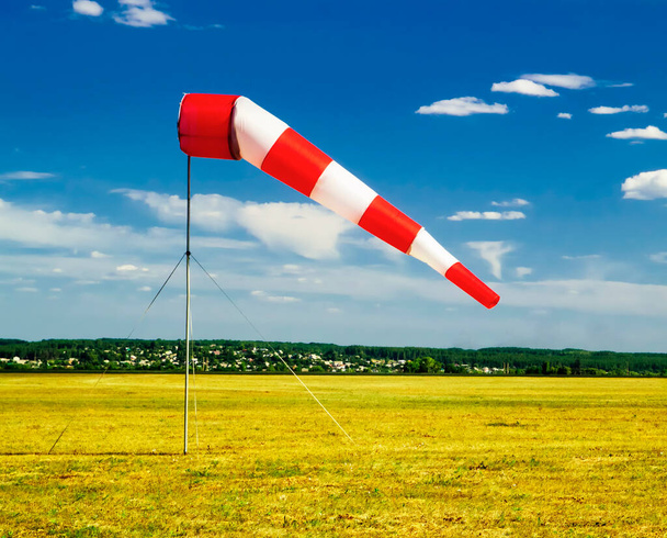 rot-weiße Windsack-Windsocke am blauen Himmel, gelbes Feld und Wolken im Hintergrund, Silhouette eines Oldtimer-Flugzeugs am Himmel - Foto, Bild