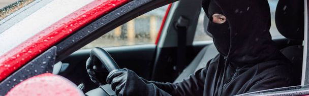 Panoramic crop of robber in balaclava sitting in auto during rain - Photo, Image