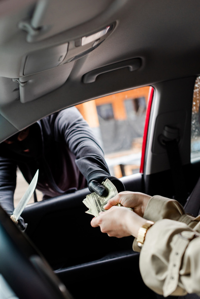 Selective focus of robber in balaclava holding knife and taking dollars from driver in car  - 写真・画像