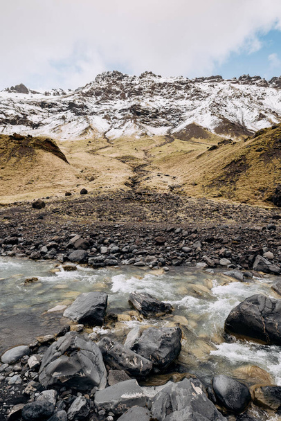 Mountain river at the foot of the mountain with a snow-capped peak. Yellow dry grass on the mountains in May in Iceland. - Фото, изображение