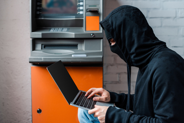 Seitenansicht des Hackers in Maske mit Laptop mit leerem Bildschirm in der Nähe des Geldautomaten  - Foto, Bild