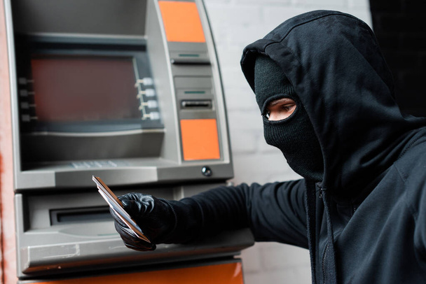 Oldalnézet a rabló símaszkban kezében hitelkártyák közelében automatizált pénztárgép - Fotó, kép