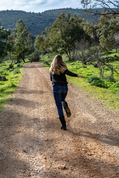 Ένα κορίτσι με φτερουγίσματα μαλλιά τρέχει κατά μήκος ενός επαρχιακού δρόμου προς την κατεύθυνση της οροσειράς - Φωτογραφία, εικόνα
