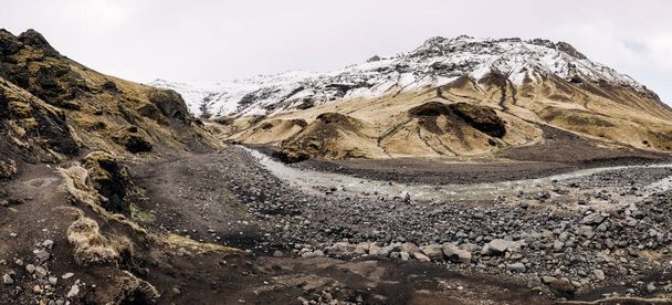 Горная река у подножия горы с заснеженной вершиной. Желтая сухая трава в горах в мае в Исландии
. - Фото, изображение