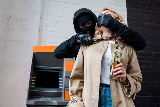 Κλέφτης με μάσκα κρατώντας μαχαίρι κοντά σε γυναίκα κρατώντας χρήματα και πιστωτική κάρτα  - Φωτογραφία, εικόνα