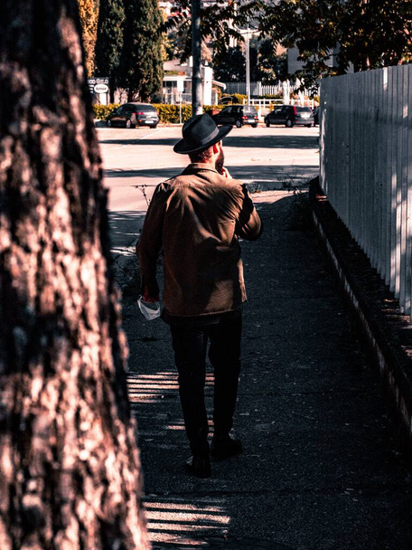 Μια κάθετη φωτογραφία ενός άνδρα με ένα μαύρο καπέλο και ένα καφέ πουκάμισο που περπατούσε στο πεζοδρόμιο την ημέρα - Φωτογραφία, εικόνα