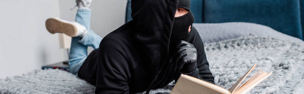 Horizontal crop of robber in balaclava holding book on bed  - Foto, imagen