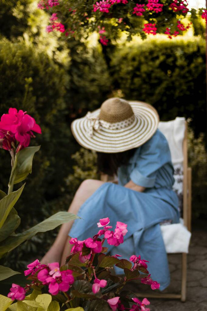 A vertical shot of a female in a straw hat sitting on a deck chair behind the pink flowers - Zdjęcie, obraz