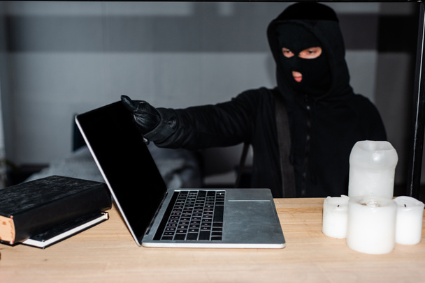 Selective focus of thief in balaclava taking laptop from table  - Foto, Imagem