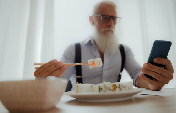 Zakenman die sushi eet tijdens de lunch en op zoek is naar een smartphone. Ondernemer in de thuisstudio. Job en modern concept - Beeld - Foto, afbeelding