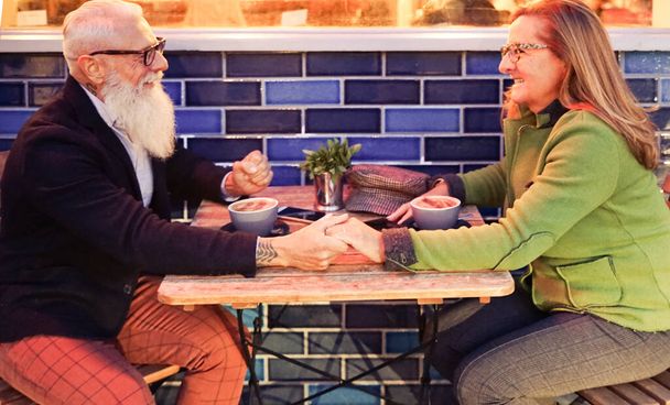 Hipster retired senior couple in love drinking hot cappuccino at outdoor cafeteria. Sweet couple, looking in their eyes. Joyful elderly lifestyle concept with wife and husband - Image - Photo, Image