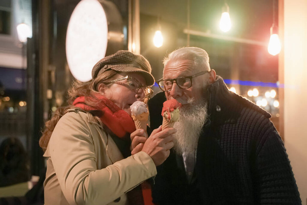Ηλικιωμένοι ζευγάρι πρόσωπο με πρόσωπο τρώει παγωτό σε στάση αγάπης. Ελκυστικοί συνταξιούχοι κοιτάζονται και διασκεδάζουν έξω από το μπαρ καφέ ζελατέρια. Έννοια της ρομαντικής και ερωτικής στιγμής - Image - Φωτογραφία, εικόνα