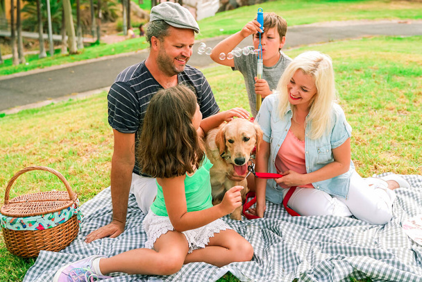 Concetto di famiglia felice - madre, padre e piccoli figli che fanno un picnic al parco estivo. Il cane è molto felice in questa famiglia. Concentrare il cane - Immagine
 - Foto, immagini