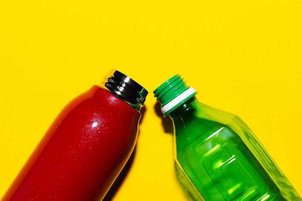 Close-up of two bottles. Steel reusable thermo water bottle and plastic disposable bottle. Isolated on yellow background. Red and green colors. - Фото, изображение