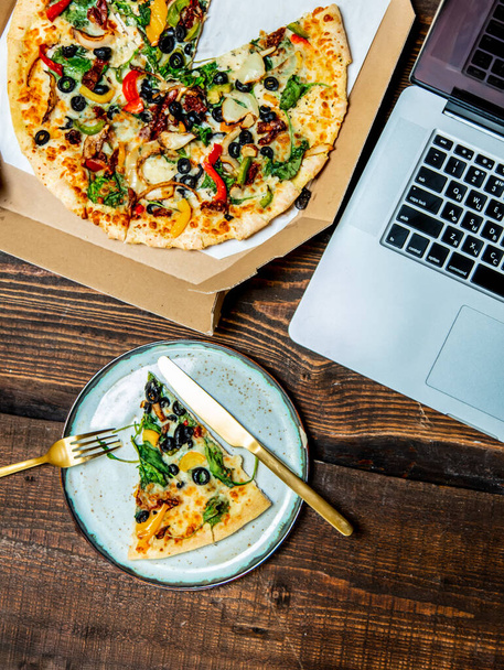 木製テーブルのラップトップコンピュータの横に段ボールでオリーブとチーズと地中海のピザ - 写真・画像