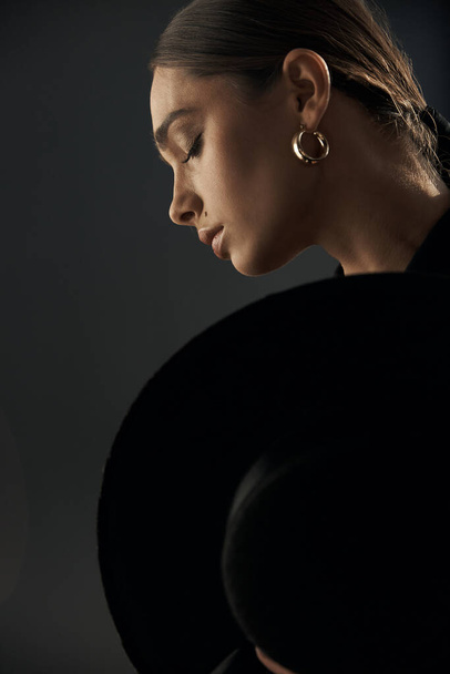 profile of a beautiful girl with closed eyes, she has a professional makeup, earrings in her ear, she and covered with a black hat on a dark background in the studio - Photo, Image