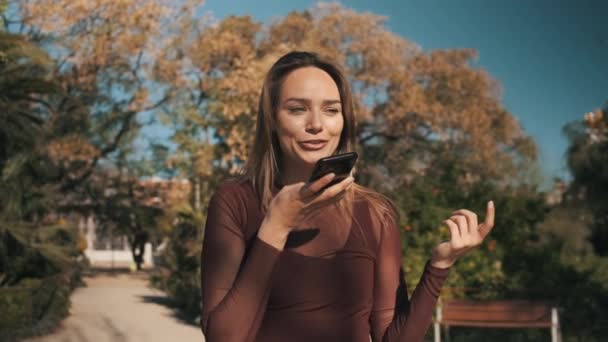 市内の公園を歩いているスマートフォンのスピーカーフォンで楽しそうに話している可愛い女の子のショットを追跡しています。陽気な美しい女の子の録音音声メッセージ屋外 - 映像、動画