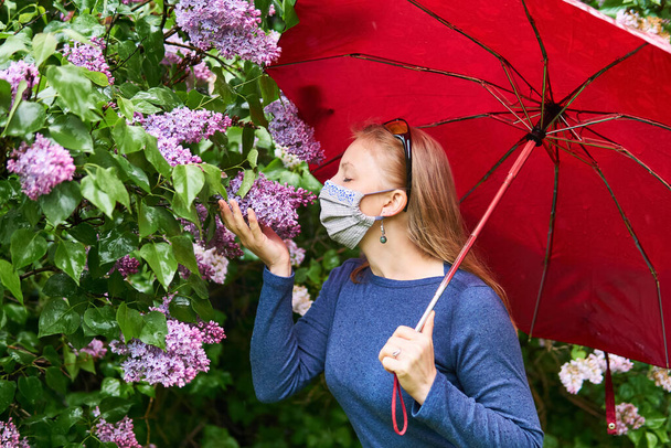 mujer en una máscara protectora casera trata de oler la inflorescencia lila en la lluvia al aire libre
 - Foto, Imagen