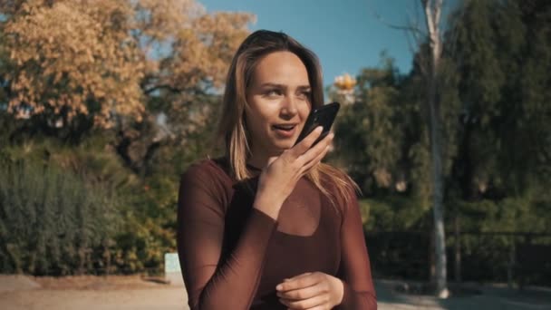 Tracking rond van mooie flirtende meisje gelukkig opnemen voice message op smartphone wandelen in het stadspark.  - Video