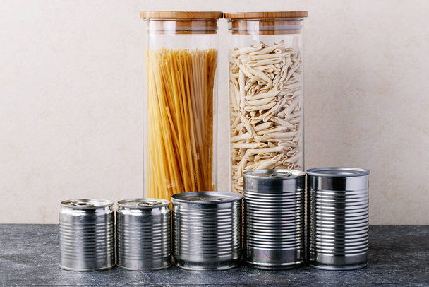 Canned food in metal cans with oats, rice, tea, and pasta in glass jars over white texture background. Copy space. Food supply concept - Foto, Imagem