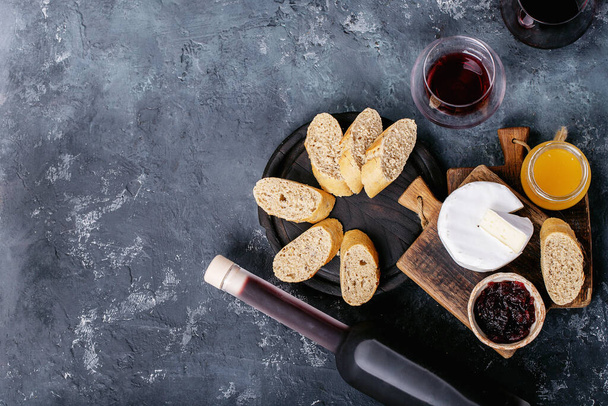 Glass of red wine served with cheese camembert, honey, jam and french baguette over dark texture background. Top view, flat lay. Copy space - Photo, Image
