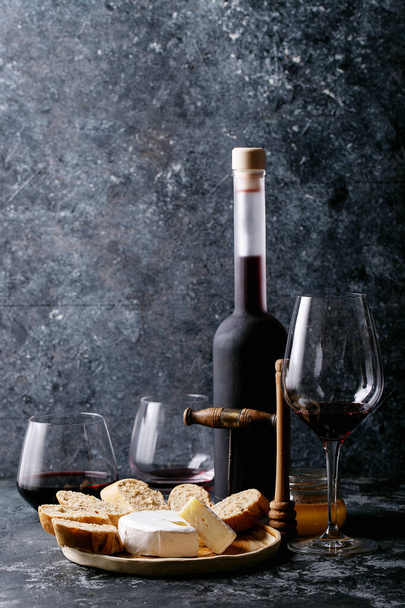 Glass of red wine served with cheese camembert, honey, jam and french baguette over dark texture background - Фото, изображение