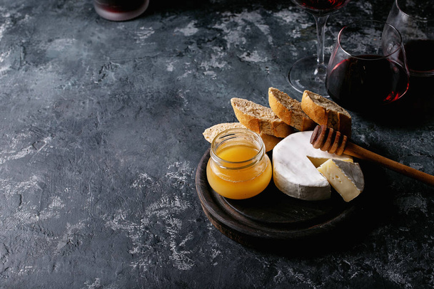 Glass of red wine served with cheese camembert, honey, jam and french baguette over dark texture background - Zdjęcie, obraz