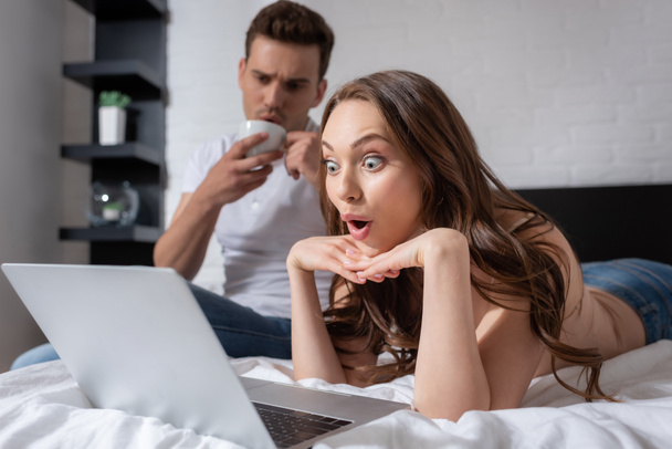 selective focus of surprised woman watching movie on laptop near boyfriend with cup  - Foto, Bild